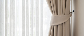 Beige fabric curtains tied back with a matching tie-back band, hanging next to sheer white vertical blinds on a window.