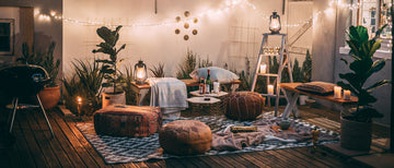 A cozy outdoor patio setup with string lights, candles, potted plants, a small table, and several poufs and blankets arranged on a wooden deck.